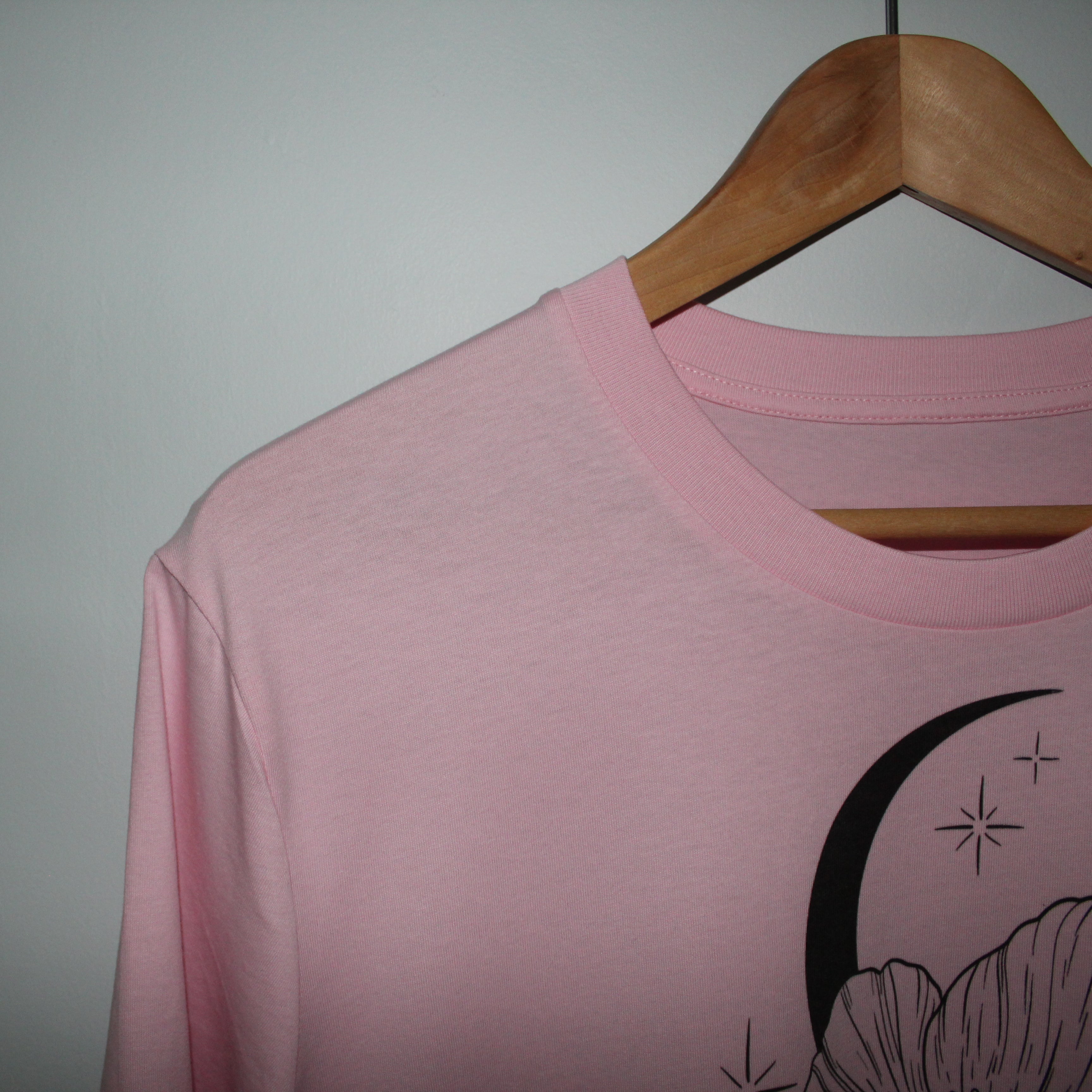 Pink t-shirt with black crescent moon and floral design on a wooden hanger against a gray background