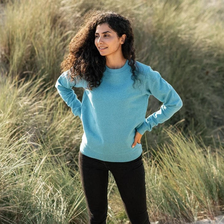Woman in a blue sweater standing in a natural setting with grass and sand.