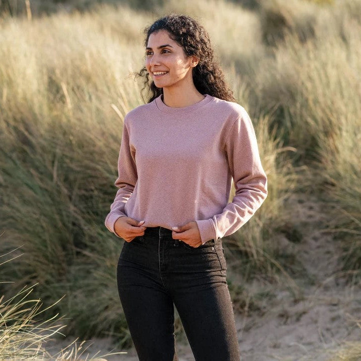 Woman standing in a sandy dune landscape wearing a pink sweater and black pants.