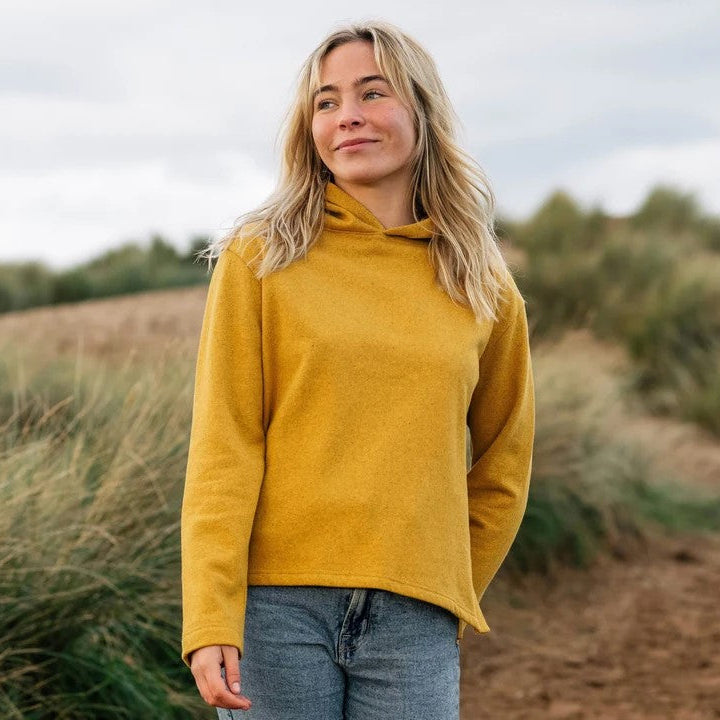 Woman wearing a yellow sweater standing in a field with grass and a cloudy sky.